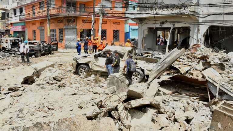 Esquina urbana con una edificación derrumbada, un vehículo sepultado y la calzada cubierta de escombros