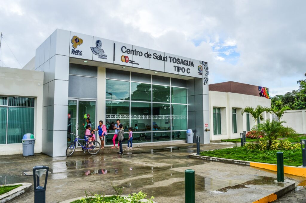 Fachada del Centro de Salud Tosagua Tipo C en funcionamiento, con rótulo del Ministerio de Salud Pública y pacientes esperando en la entrada acristalada