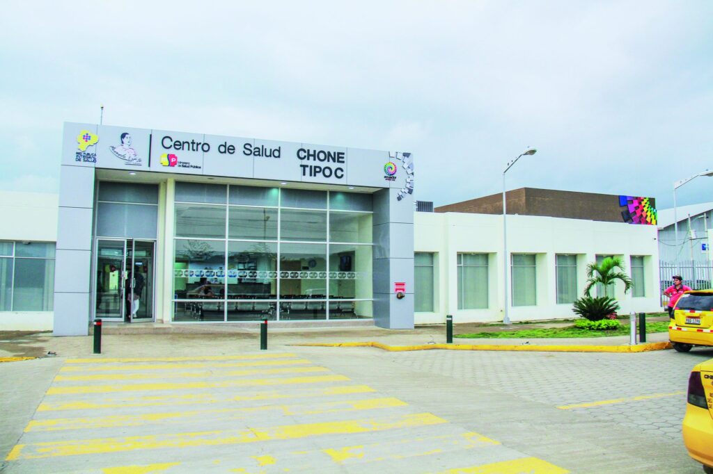 Fachada del Centro de Salud Chone Tipo C reparado y en funcionamiento, con sala de espera visible tras el ventanal y taxis en el parqueadero