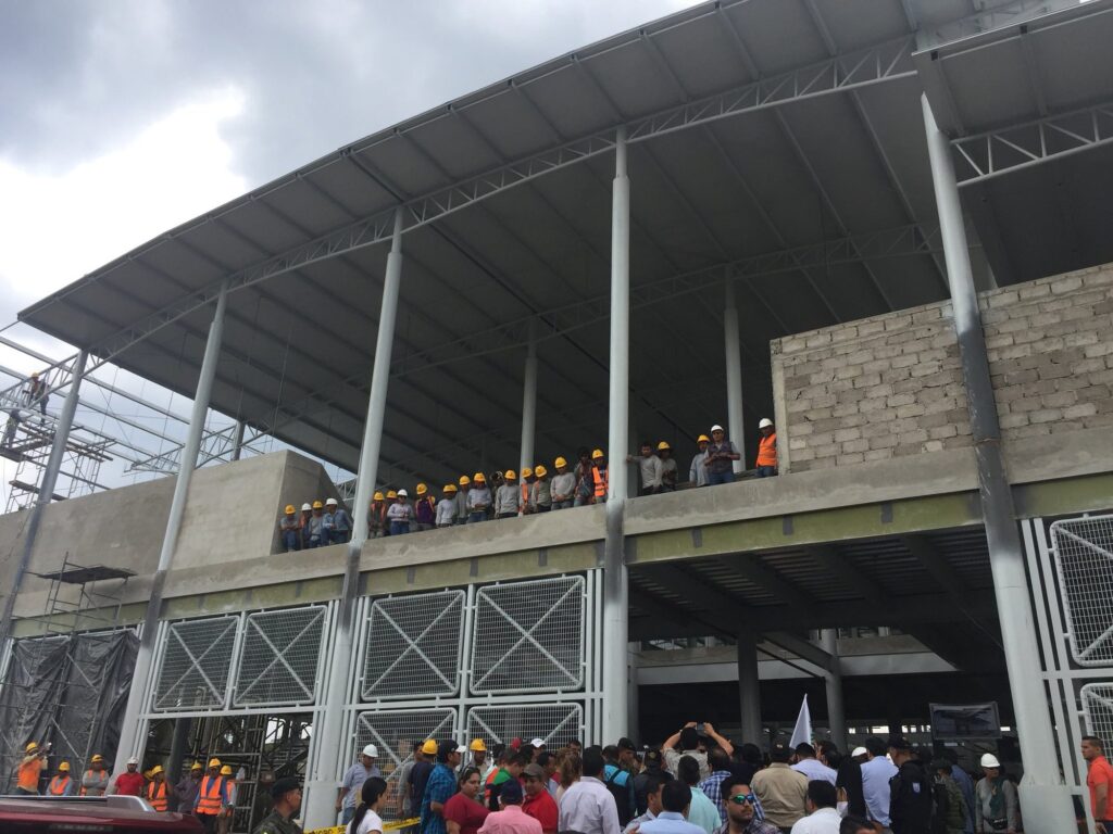 Estructura metálica del mercado de Calderón en construcción, con obreros con casco sobre la losa y autoridades reunidas en el ingreso