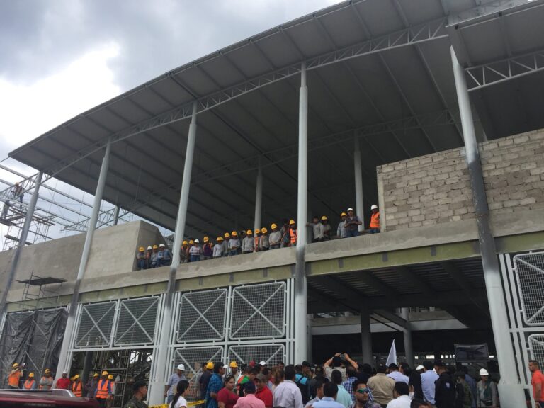 Estructura metálica del mercado de Calderón en construcción, con obreros con casco sobre la losa y autoridades reunidas en el ingreso