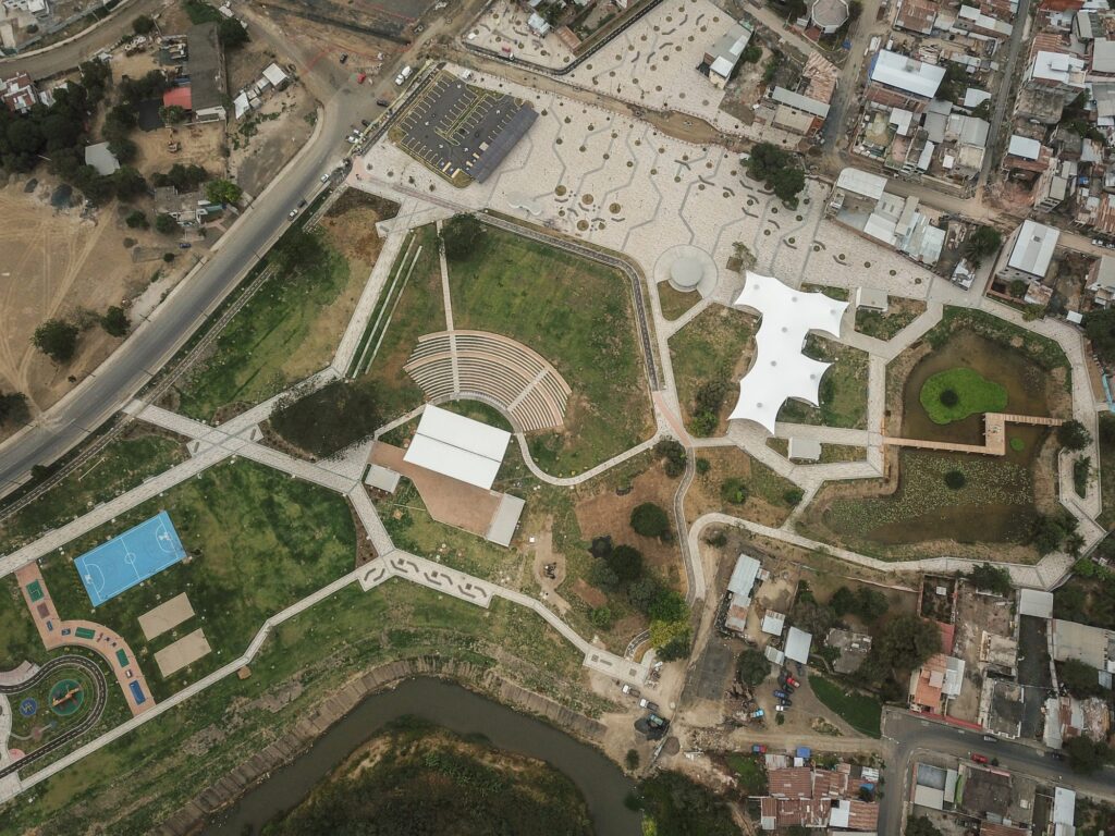 Vista aérea cenital del Parque Las Vegas de Portoviejo en funcionamiento: anfiteatro, canchas deportivas, juegos infantiles, laguna con pasarela y plaza de acceso