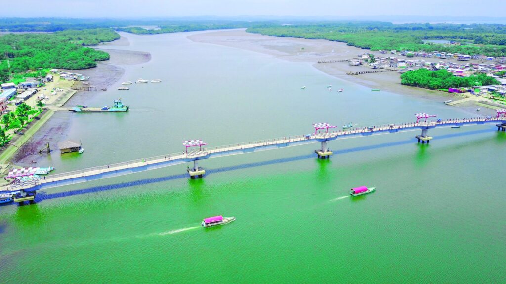 Vista aérea del puente de Muisne sobre el estuario, con paso peatonal, miradores techados y embarcaciones navegando bajo la estructura
